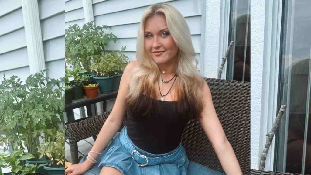 Nikki Wilde sits on a wicker balcony chair, wearing a black tank top and denim skirt with belt, layered necklaces, with potted plants nearby.