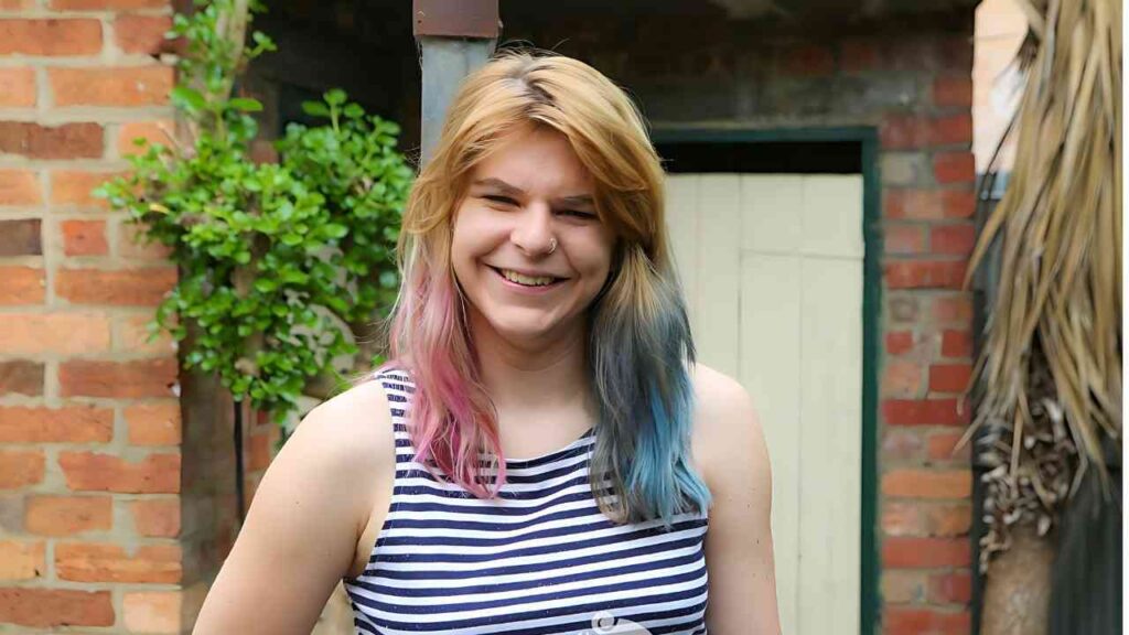 Ophelia Rose with blonde hair and pink- and blue-tipped ends, wearing a navy-and-white striped tank, stands beside brick wall.