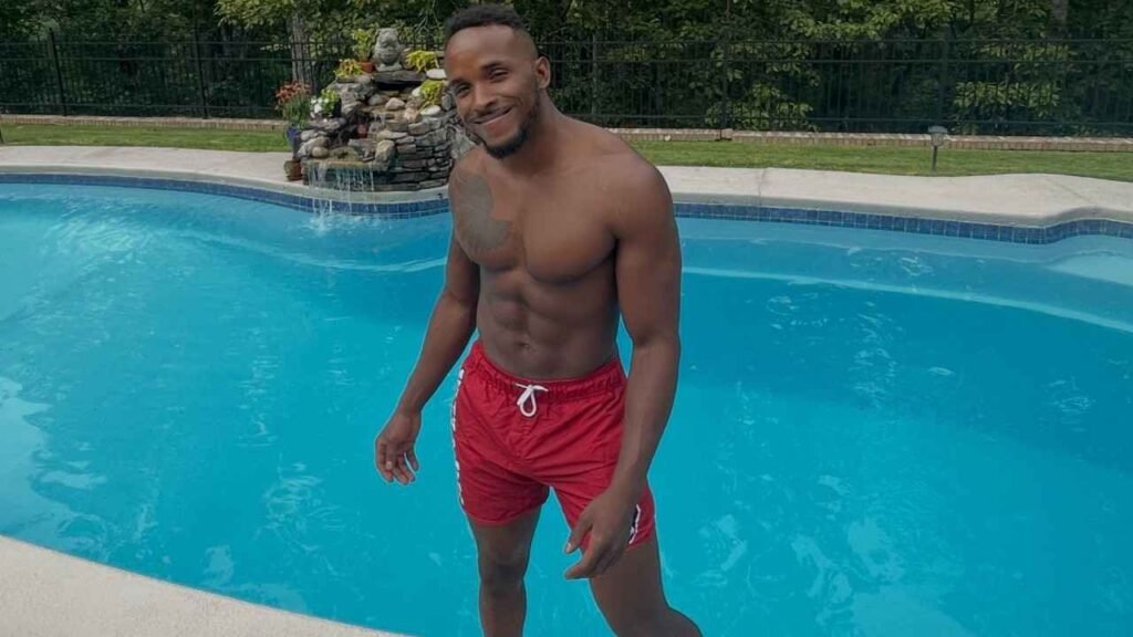 Pheonix Fellington in red swim trunks stands at the pool edge, blue water and a stone waterfall behind him.