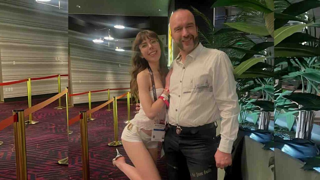 Randy Denmark in a light striped shirt stands beside a kneeling woman in white, amid red rope barriers and lush plants.