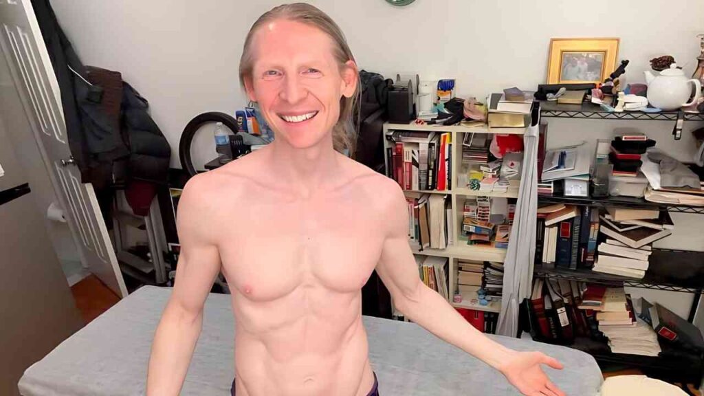 Rob Yaeger with shoulder-length blond hair stands in a cluttered room, arms out, book-filled shelves behind.