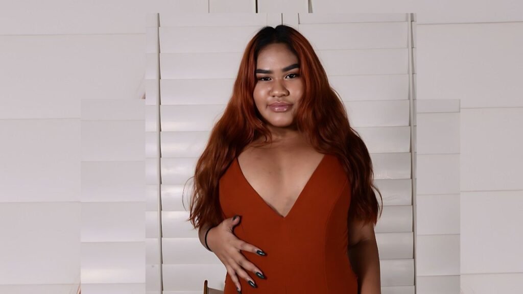 Rozey Royalty with long auburn hair in a rust V-neck dress stands before a white backdrop, hand on her abdomen, dark green nails.