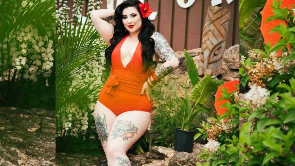Ruby Valentine in an orange swimsuit with tattoos stands outdoors among tropical plants, posing with one hand on her hip and a red flower in her hair.