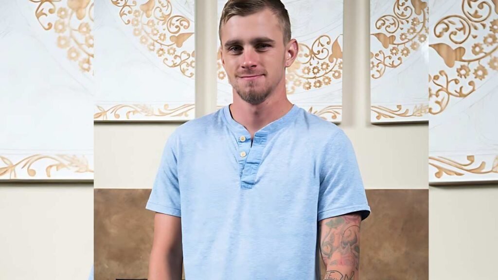 Ryan Jordan with short hair and a light beard in a Henley shirt, tattooed left forearm, smiling in front of cream walls with gold floral panels.