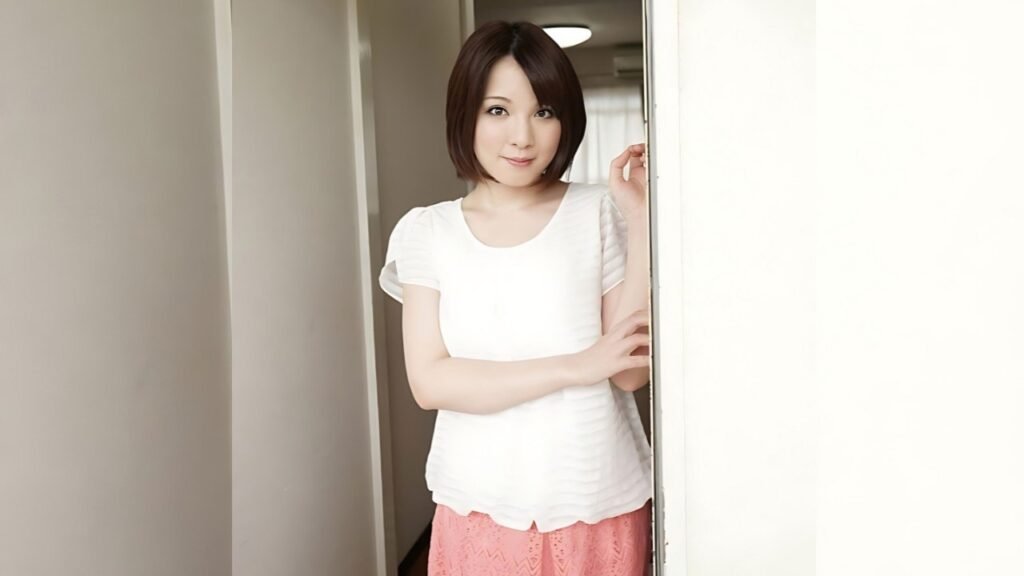 Ryo Tsujimoto with a short dark bob stands in a doorway, one hand on the frame, wearing a white top and pink skirt