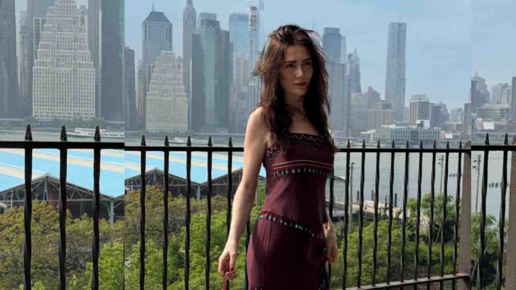 Sara Belle with dark hair in a burgundy dress with turquoise bead trim stands at a balcony railing, city skyline and river behind.