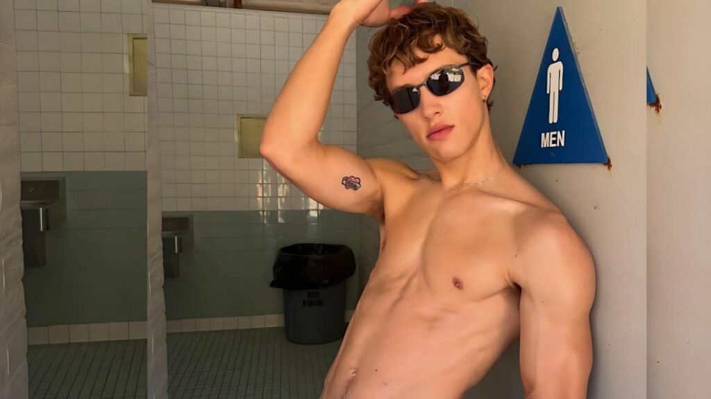 Sean Ford with curly hair and sunglasses leans against a tiled restroom wall, arm up, beside a blue MEN sign.