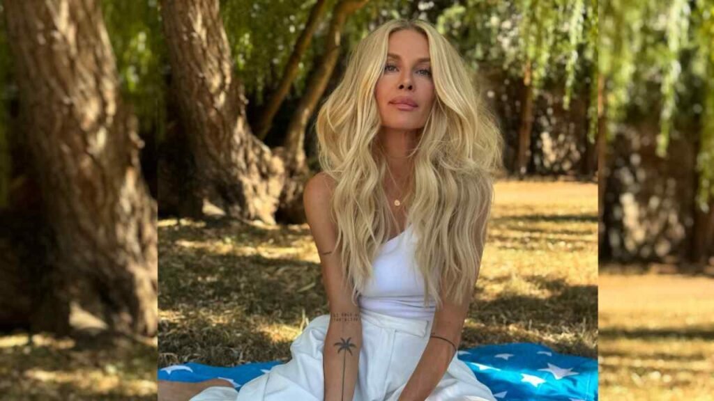 Simona Krainova with long wavy hair sits on a blue star-pattern blanket in a sunlit park, wearing a white one-shoulder dress.