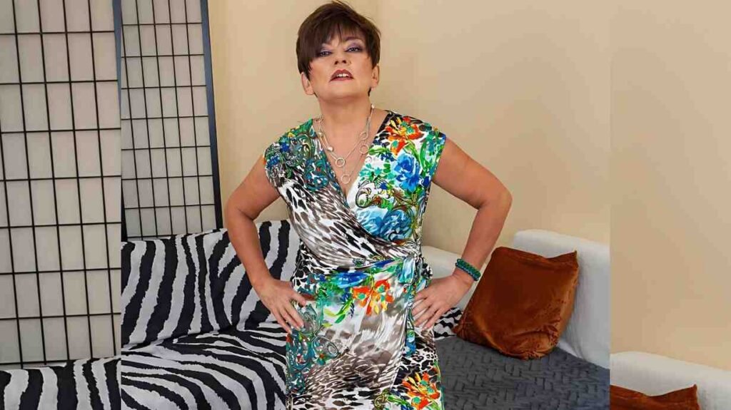 Sisi Sinaloa Short-haired, bold-makeup figure wears a bright floral wrap dress, hands on hips in front of a zebra-print sofa and wall.