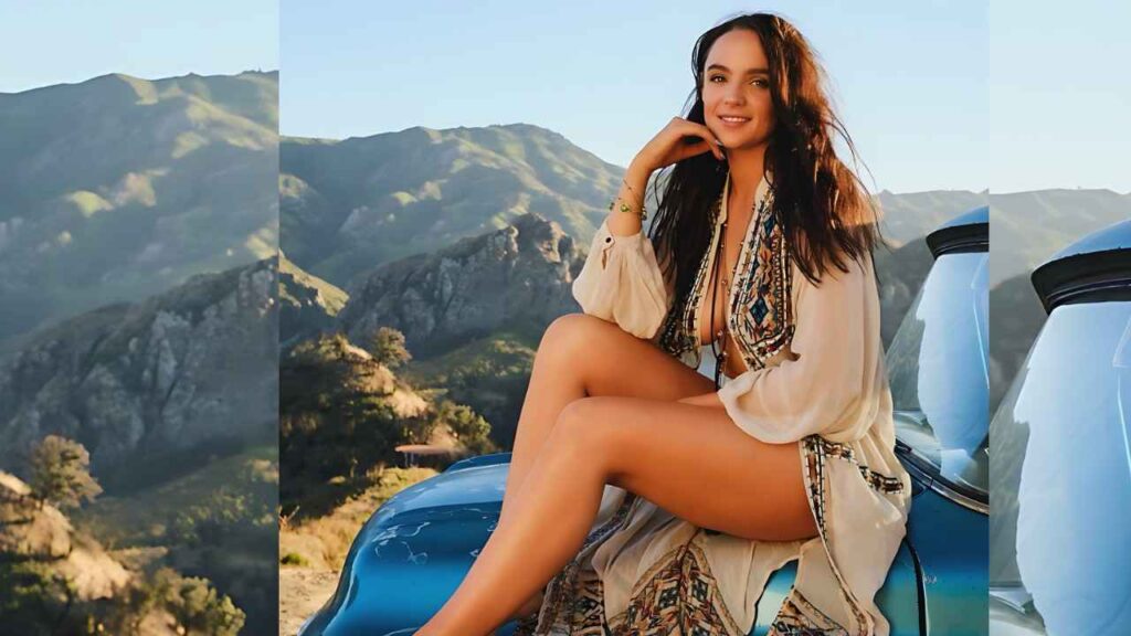 Stevie Lynn in a flowing beige embroidered dress sits on a blue vintage car against sunlit mountains and blue sky.