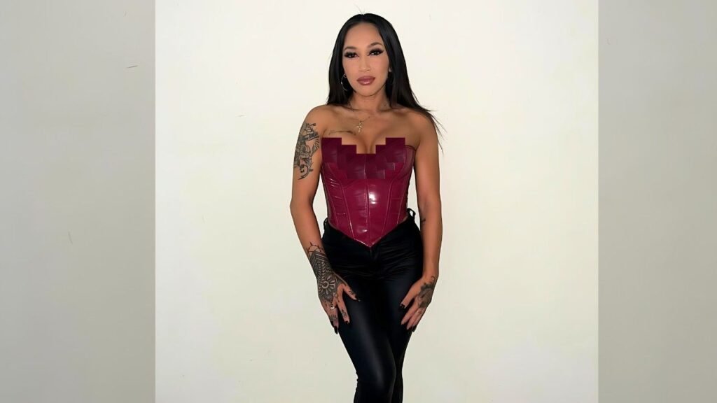 Summer Daize with long dark hair wearing a burgundy strapless corset and black shiny pants, tattoos on her arms, standing.