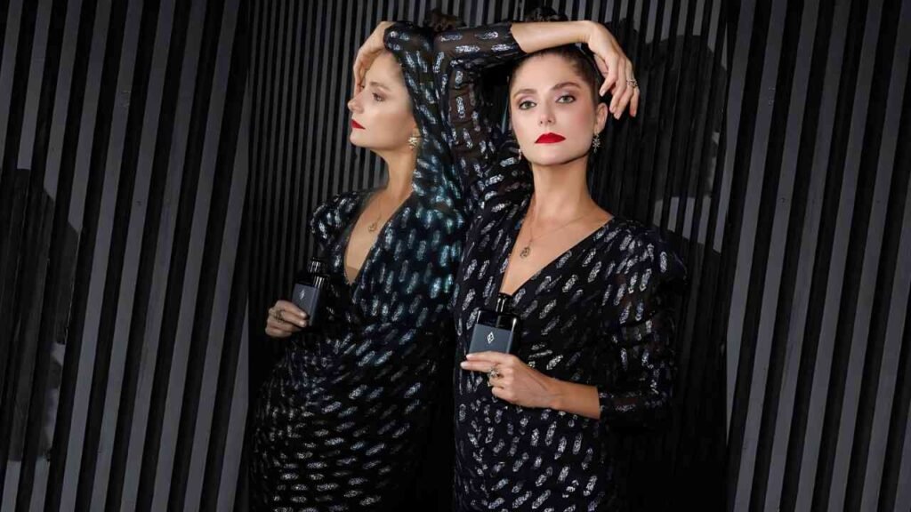 Tammy Di Calafiori in black dresses with silver patterns pose against a dark striped wall; one arm raised, both hold perfume bottles.