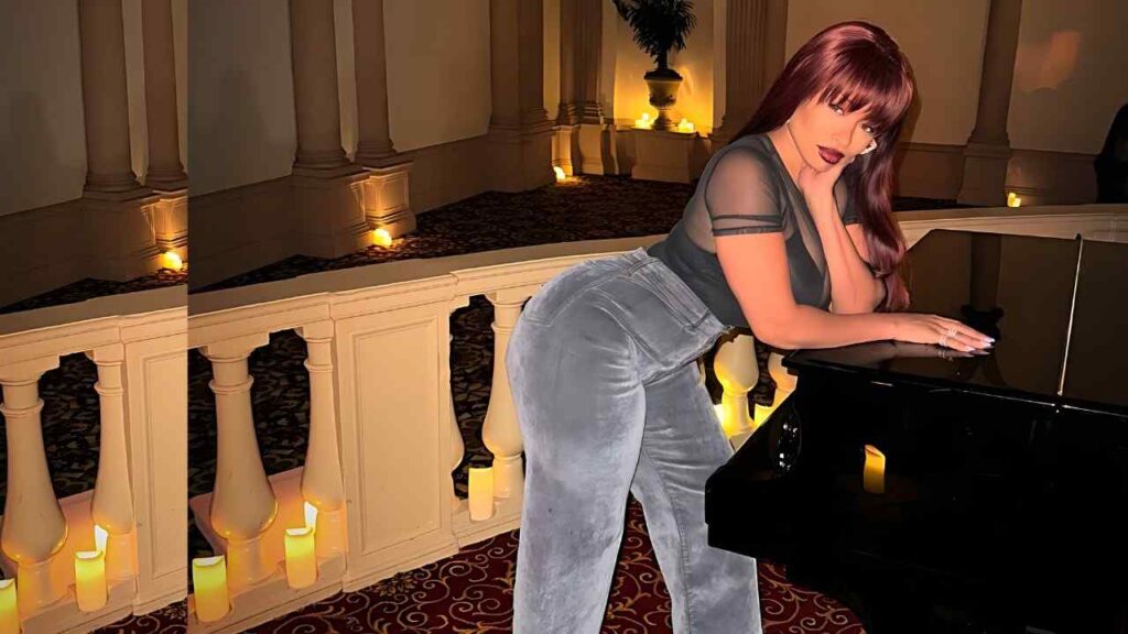 Teresa Celeste leans on a glossy black piano in a candlelit, columned hallway, wearing a sheer top and gray velvet pants.