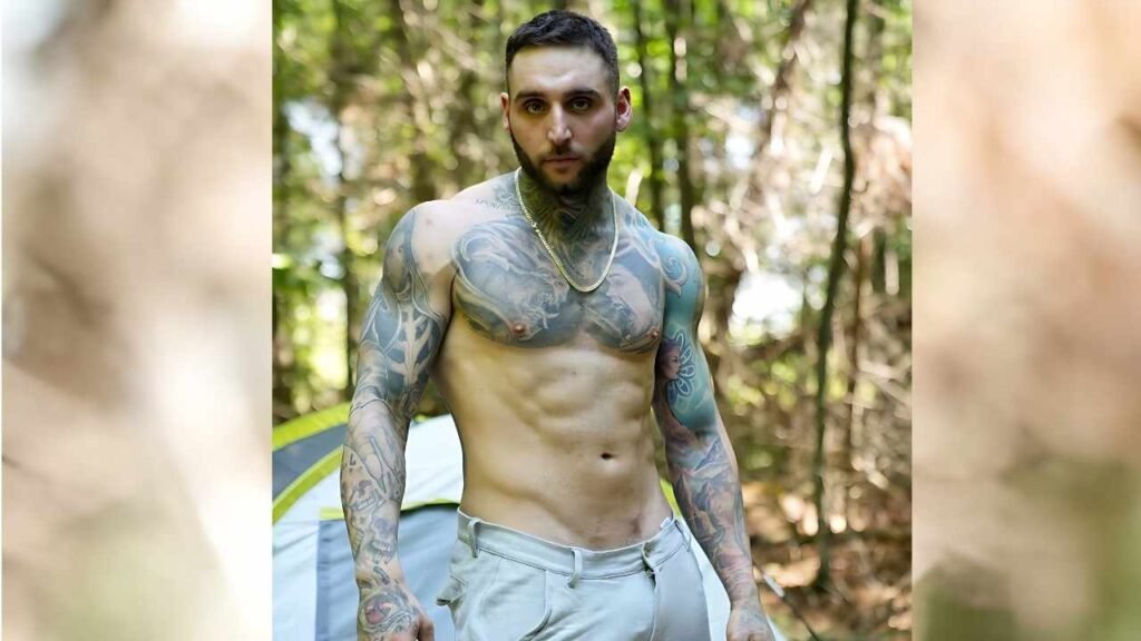 Tony Dangelo with blue-gray tattoos stands in a sunlit forest, wearing a gold chain and light shorts, tent behind.