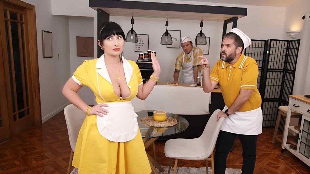 Chad Alva in a yellow retro dress and white apron strikes a pose in a kitchen set, while two chefs argue in the background.