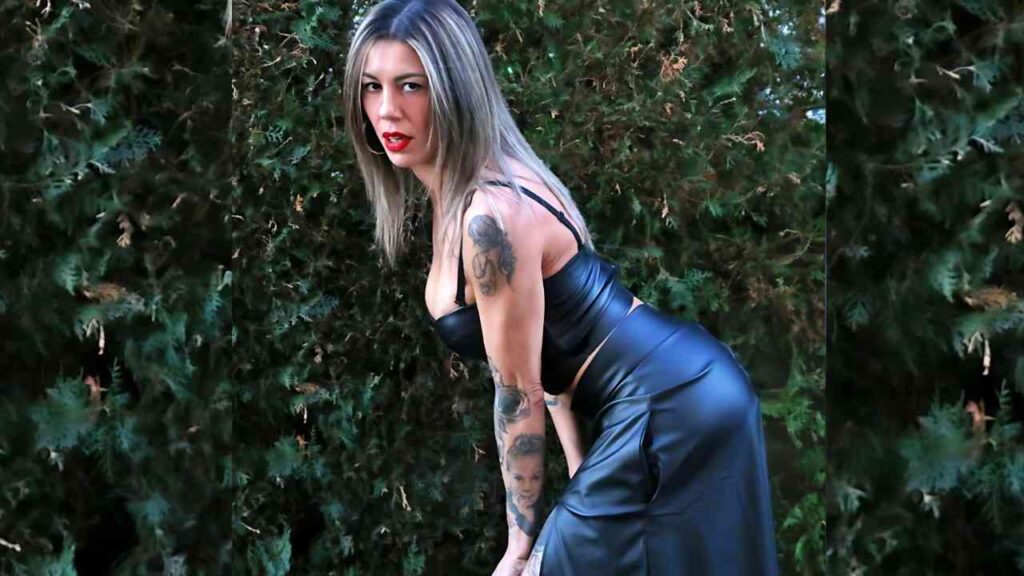 Valeria Curtis in a tight black leather dress bends forward outdoors, red lipstick, blonde hair, green hedge behind.