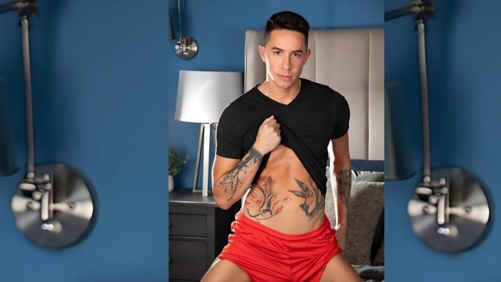 Vincent Oreilly in a black tee and red shorts lifts his shirt to show tattooed abs and arms, in a blue bedroom with a bed and lamp.