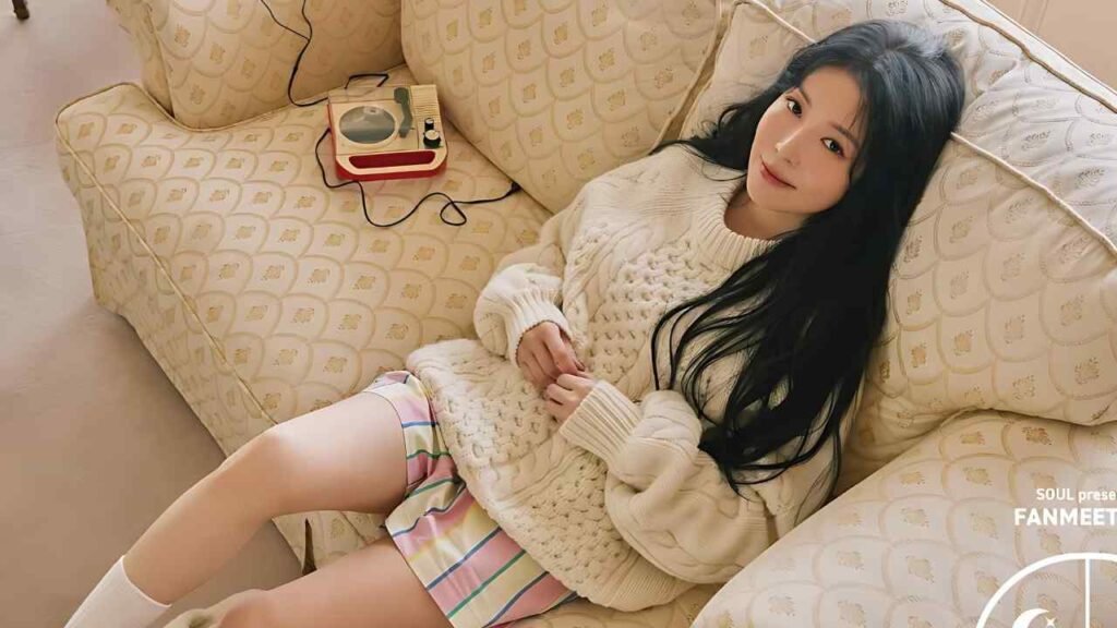 BoA lounges on a cream sofa, wearing a chunky white knit sweater and pastel striped shorts, smiling softly.