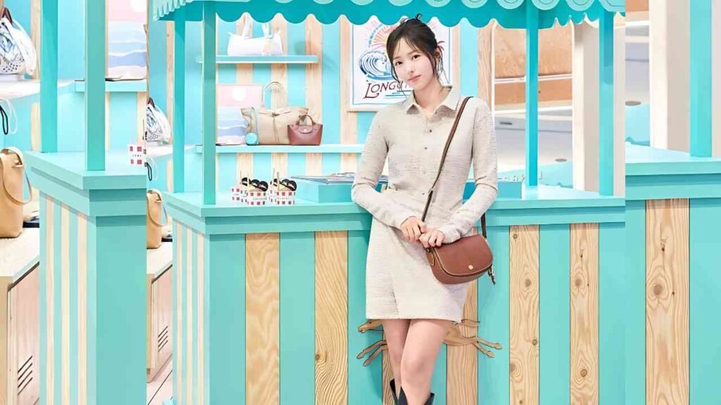 Chae Soo-bin in a cream knit dress leans on a turquoise counter in a pastel shop, brown crossbody bag slung over her.