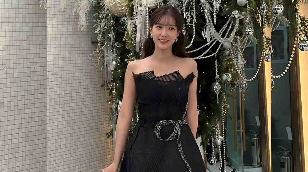 Im Soo-hyang ( 임수향 ) in a strapless black dress with jagged neckline and belt with round buckle stands before silver bead garlands, tiled wall left.