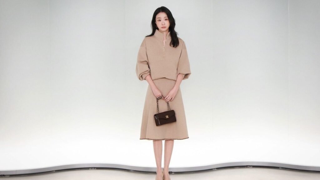Kim Da-mi stands in a studio wearing a beige knit two-piece with oversized sleeves, holding a small dark brown handbag.