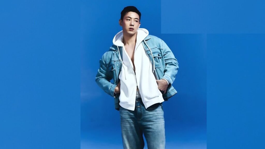 Kim Ji-soo in a light denim jacket over a white hoodie, blue jeans, hands in pockets against a vivid blue backdrop.