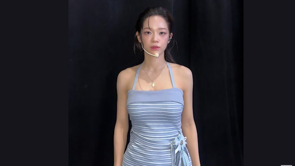 Kim Sejeong in a light blue, striped ruched halter dress stands before a dark backdrop, wearing a headset mic and a necklace.