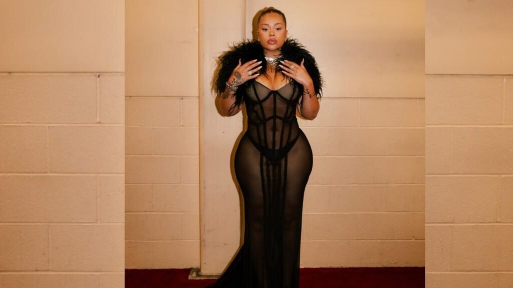 Latto in a sheer black, floor-length mesh gown with cage-like panels and a feathered boa, in a beige hallway.