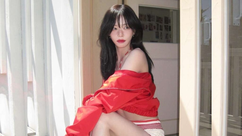 Lee Chae-young with long black hair and red lipstick sits by a sunlit doorway, wearing a red off-shoulder jacket and white shorts, gazing sideways.
