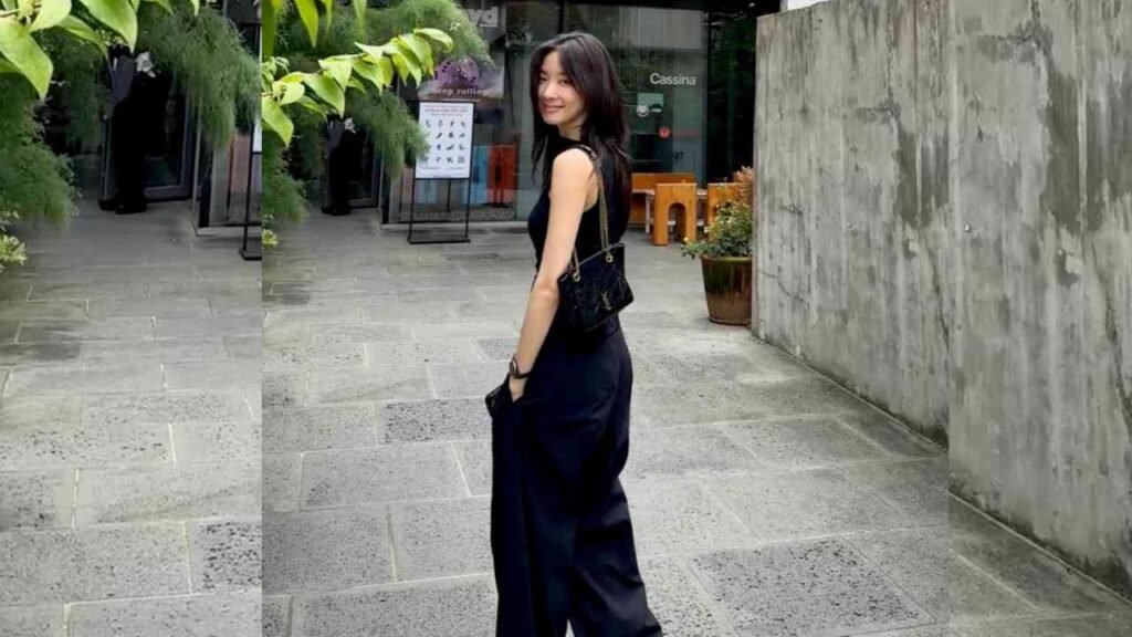 Lee Chung-ah in a sleeveless black top and wide-leg pants poses sideways, carrying a small black handbag on a stone plaza.