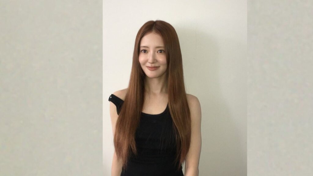 Lee Se-young ( 이세영 ) with long, straight brown hair and a black off-shoulder top stands against a pale wall, smiling softly.