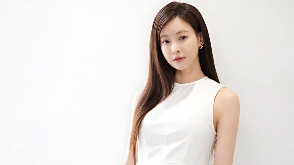 Oh Yeon-seo ( 오연서 ) with long straight brown hair wearing a white sleeveless top stands against a pale wall, gazing softly at the camera.