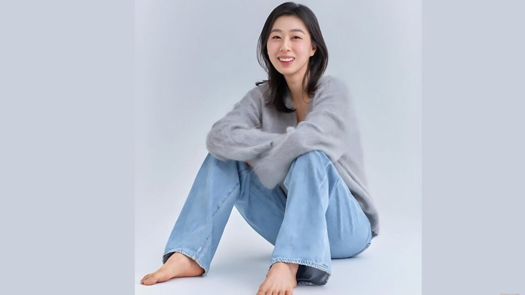 Park Ji-yeon sits cross-legged on a pale studio floor, wearing a soft gray sweater and light blue jeans, smiling warmly with arms around her knees.