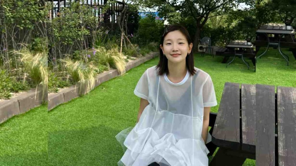 Park So-dam in a pale sheer white dress sits on a dark wooden bench in a sunny garden, smiling softly amid lush greenery.