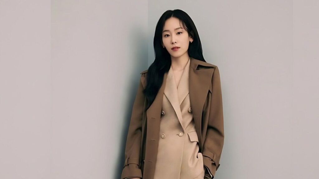 Seo Hyun-jin with long black hair leans against a gray wall, wearing a tan trench coat over a beige suit, calm expression.