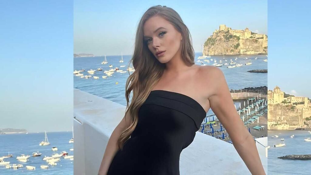 Abigail Cowen in a black dress poses gracefully by the water's edge, with a serene landscape in the background.