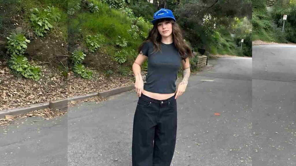 Acacia Brinley in a stylish hat and black pants poses on a bustling street, showcasing her fashion against an urban backdrop.