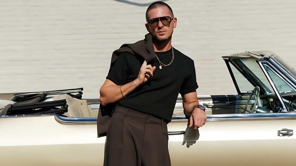 Adam Senn wearing sunglasses leans casually against a vintage car, showcasing a relaxed and stylish demeanor.