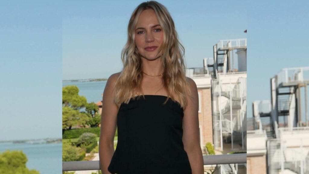 Adelaide Clemens in a black dress stands elegantly on a balcony, overlooking a scenic view.