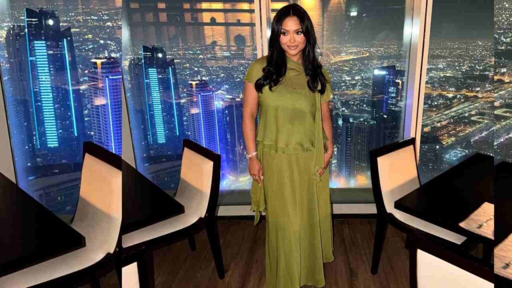 Afshan Azad in olive-green dress stands in a chic restaurant by a window, with a city skyline glowing at night outside.