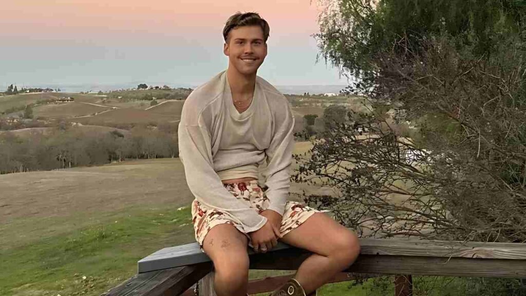 Aidan Alexander sits on a bench with his legs crossed, appearing relaxed and contemplative in a park setting.