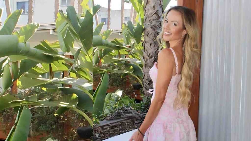 Amanda Carraway with wavy blonde hair in a pink dress stands beside a corrugated metal wall as tropical plants glow in sun.