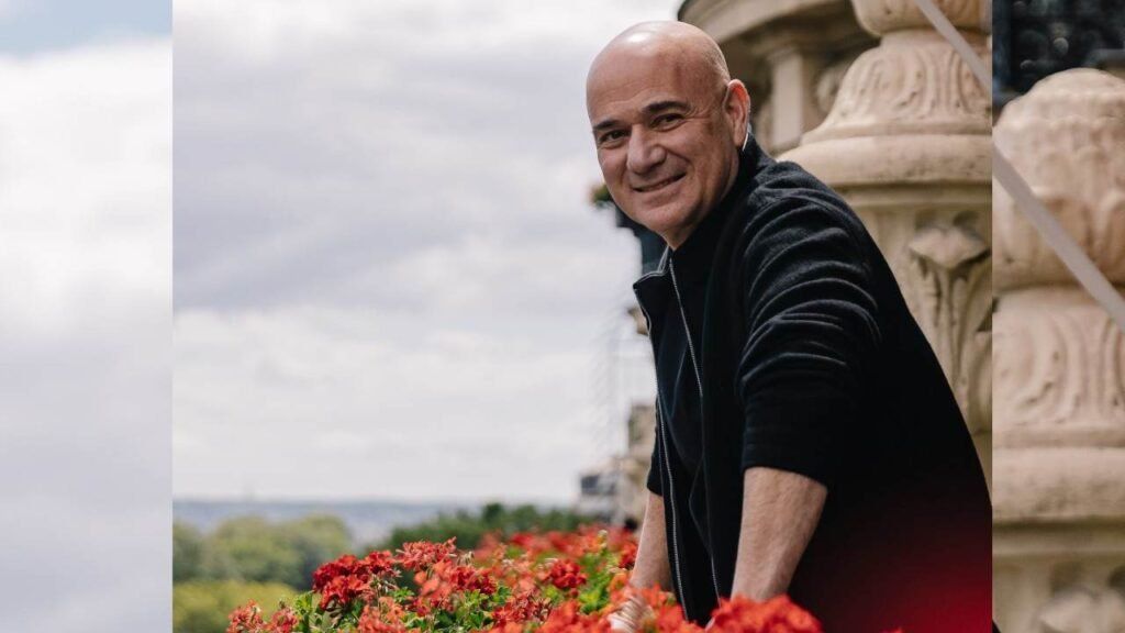 Andre Agassi in a black jacket leans on a railing beside carved stonework, with red flowers in the foreground.