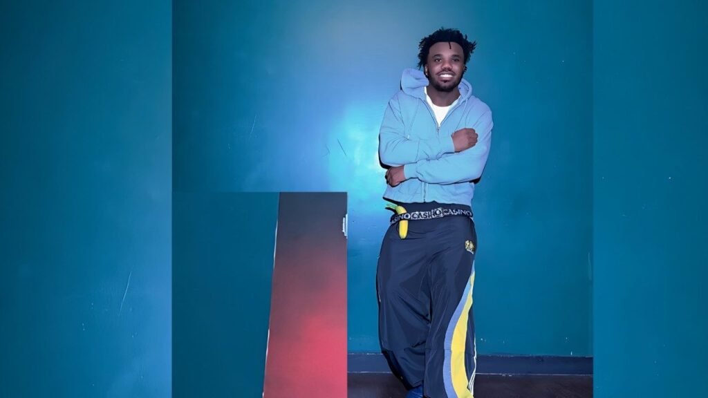 Baby Keem in a light blue hoodie and white tee, arms crossed in a teal room beside a red panel; navy pants, yellow stripes.