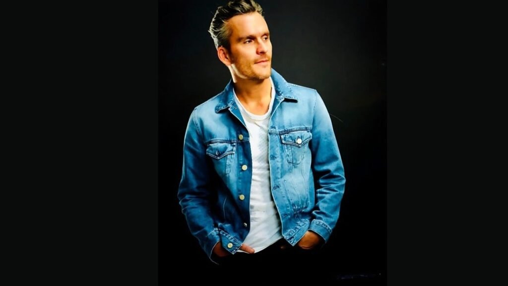 Balthazar Getty wearing a denim jacket smiles while posing for a photo against a neutral background.