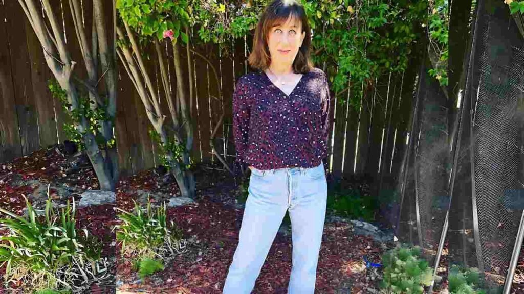 Beth Hall in a blouse and jeans stands near a fence, appearing relaxed and enjoying the outdoor environment