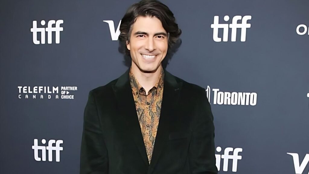 Brandon Routh in a green suit smiles confidently while standing on the red carpet at a glamorous event.