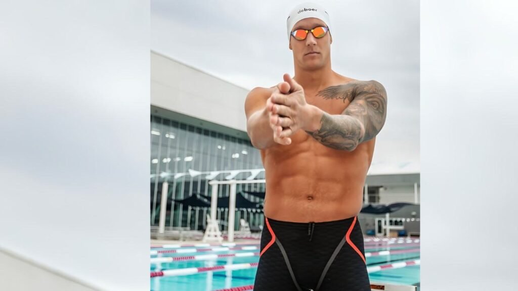 Caeleb Dressel swimmer in white cap and orange mirrored goggles stretches toward the camera, tattooed arm visible beside the pool.