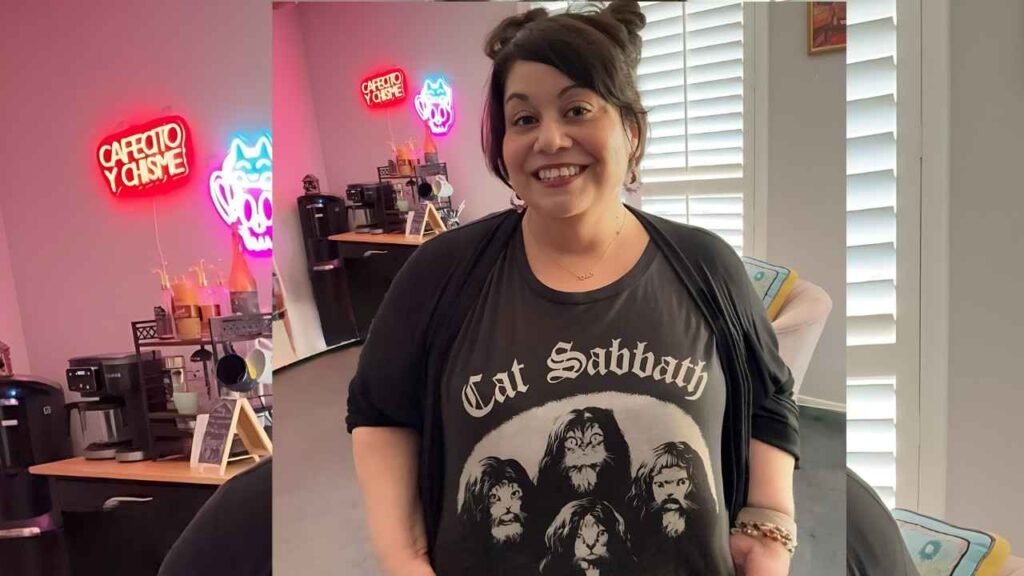 Carla Jimenez in a black cardigan and Cat Sabbath t-shirt stands in a cafe with neon cat signs behind her.