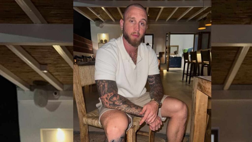 Chet Hanks with tattoo sleeves sits on a wooden chair at a kitchen counter in a modern open-plan room, wearing a white polo.